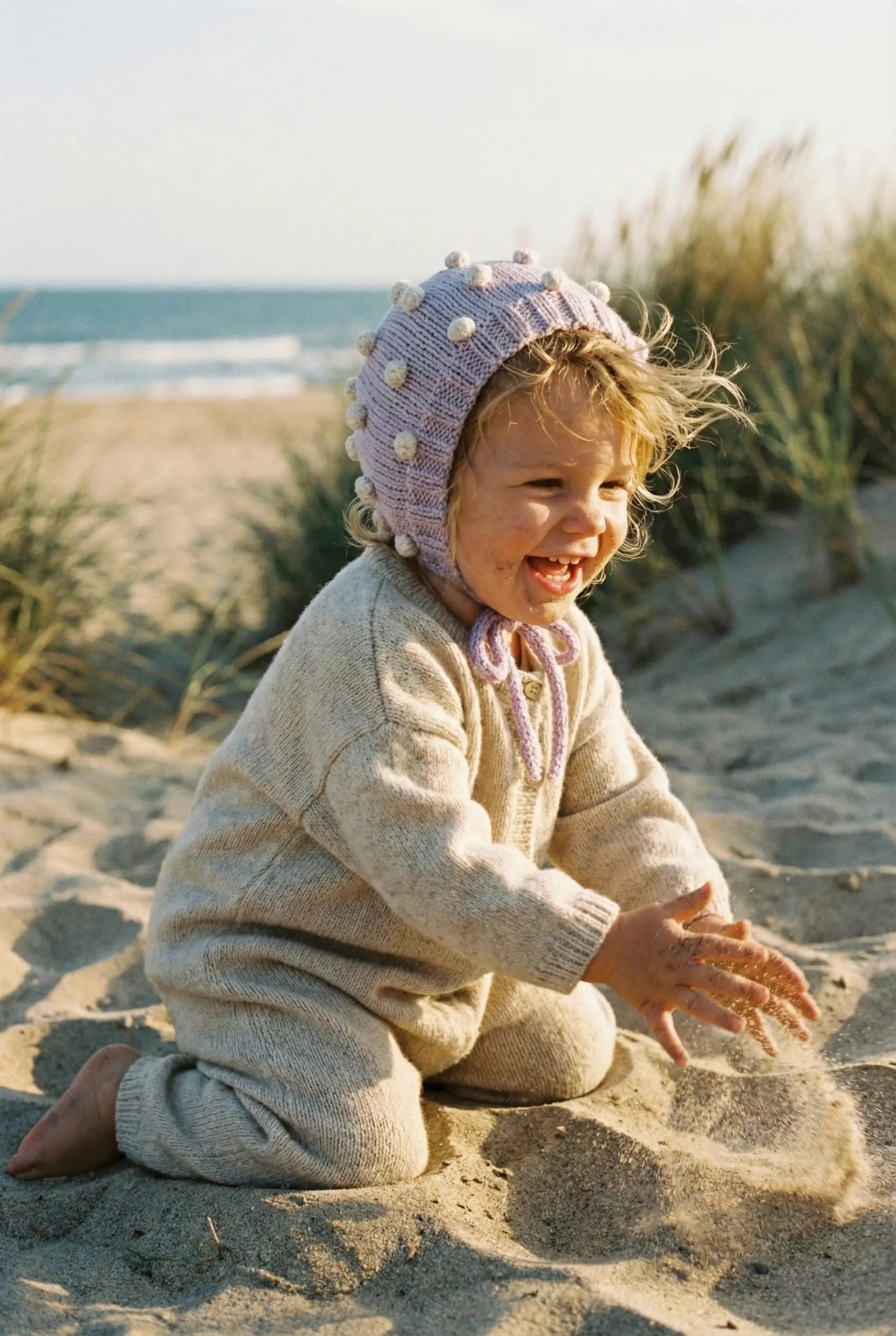 Baby wearing a lavender organic cotton bonnet playing with sand on a beach