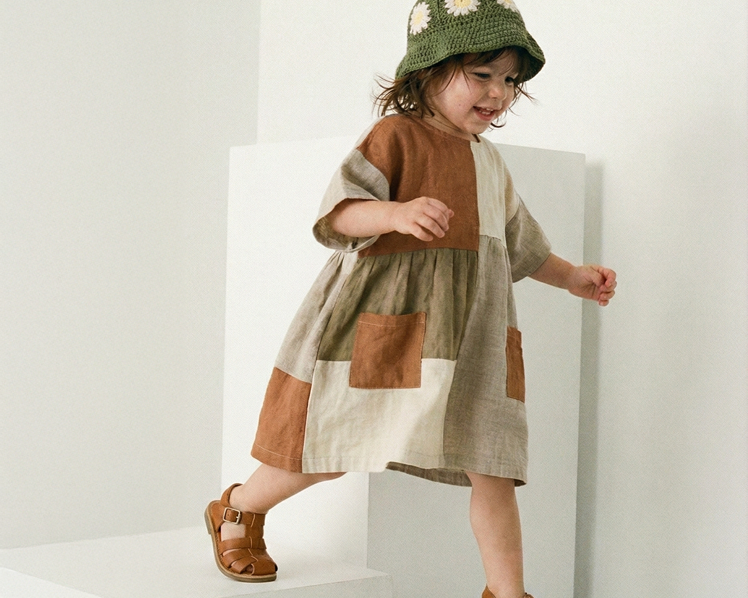 Dziecko w szydełkowym zielonym kapeluszu dziecięcym Daisy i wzorzystej sukience na białych schodkach | Child wearing a patterned dress and crocheted deep green summer hat on white steps