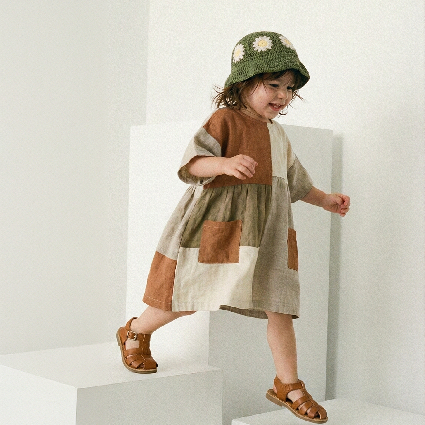 Dziecko w szydełkowym zielonym kapeluszu dziecięcym Daisy i wzorzystej sukience na białych schodkach | Child wearing a patterned dress and crocheted deep green summer hat on white steps