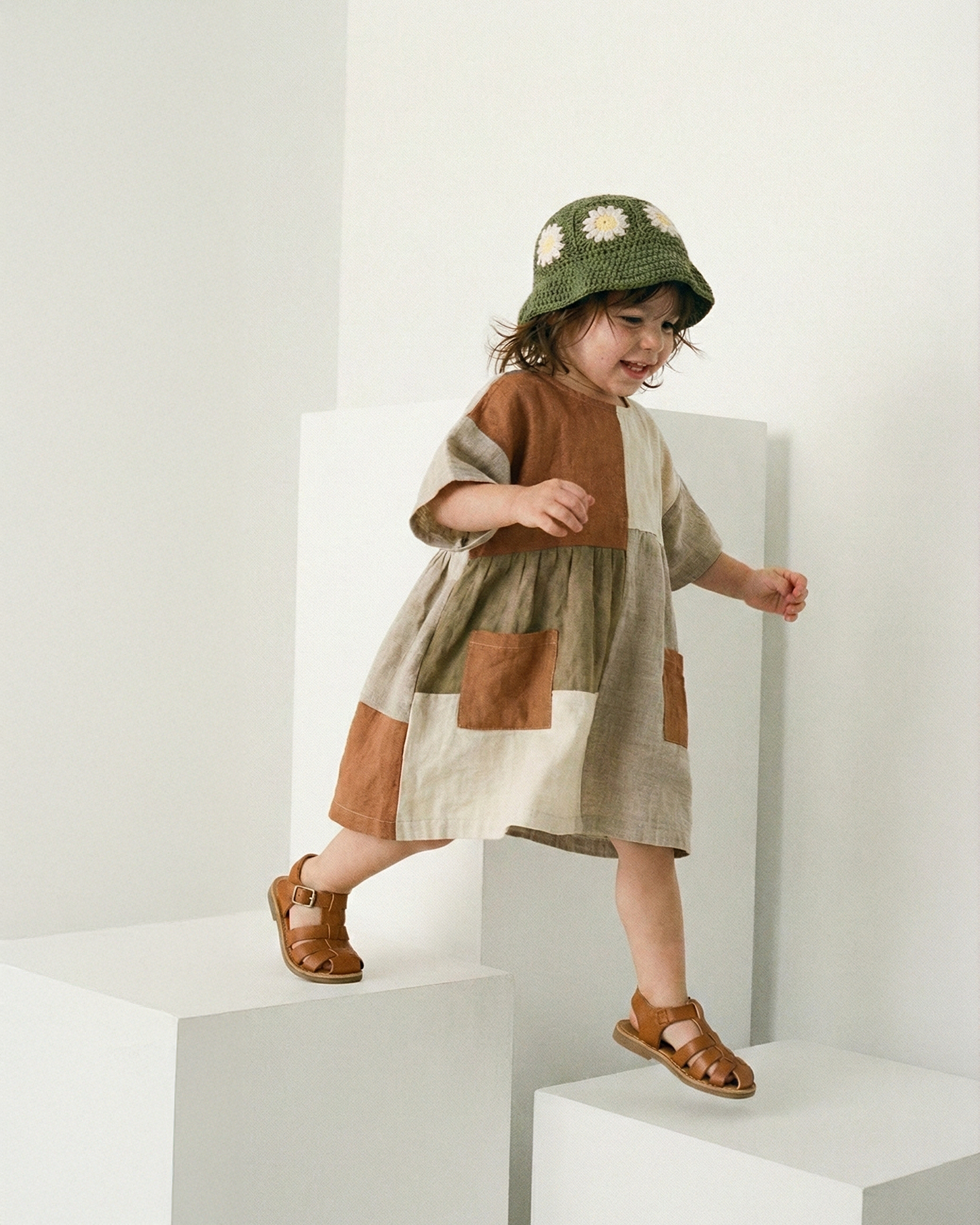 Dziecko w szydełkowym zielonym kapeluszu dziecięcym Daisy i wzorzystej sukience na białych schodkach | Child wearing a patterned dress and crocheted deep green summer hat on white steps
