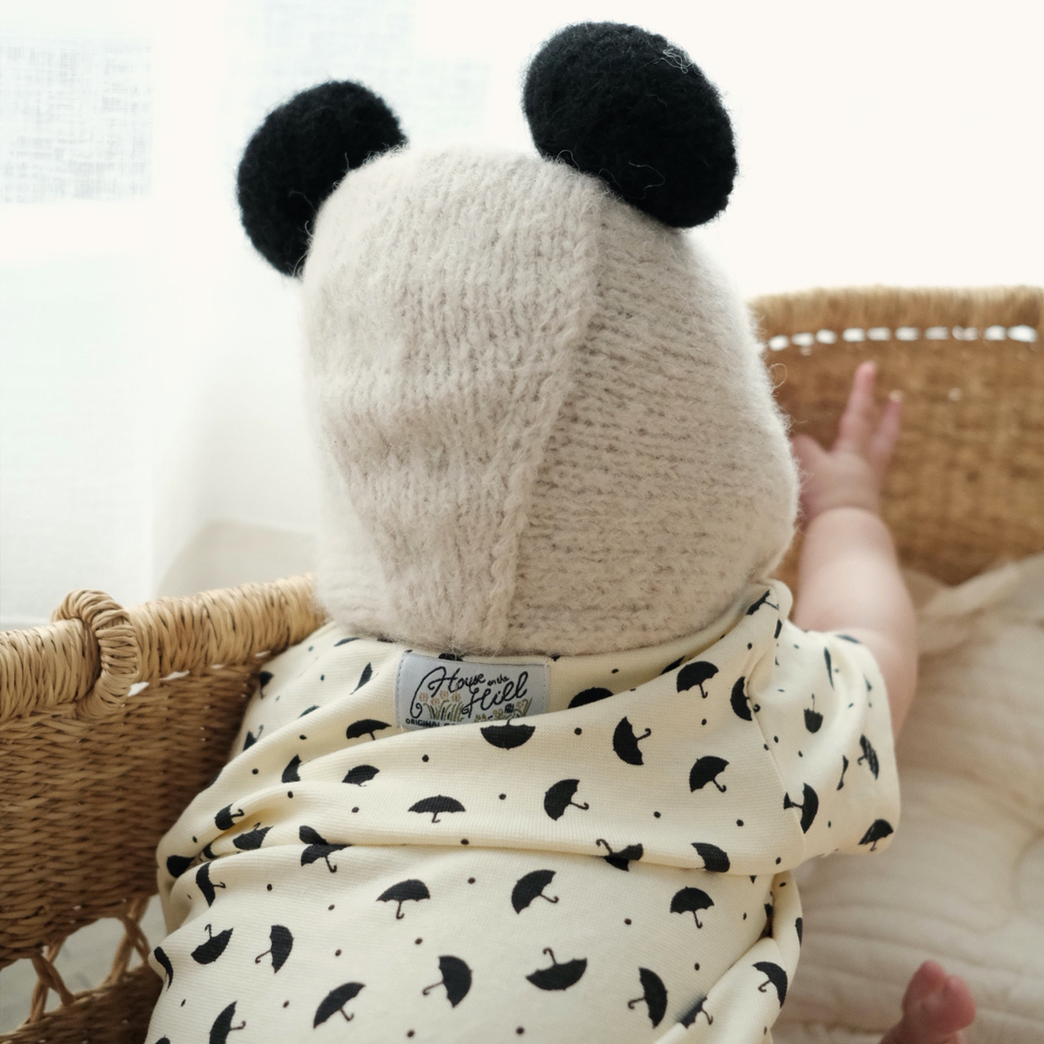 A baby seen from behind wearing a cream knit panda-ear bonnet and a cream onesie with a black umbrella print, sitting in a wicker basket.