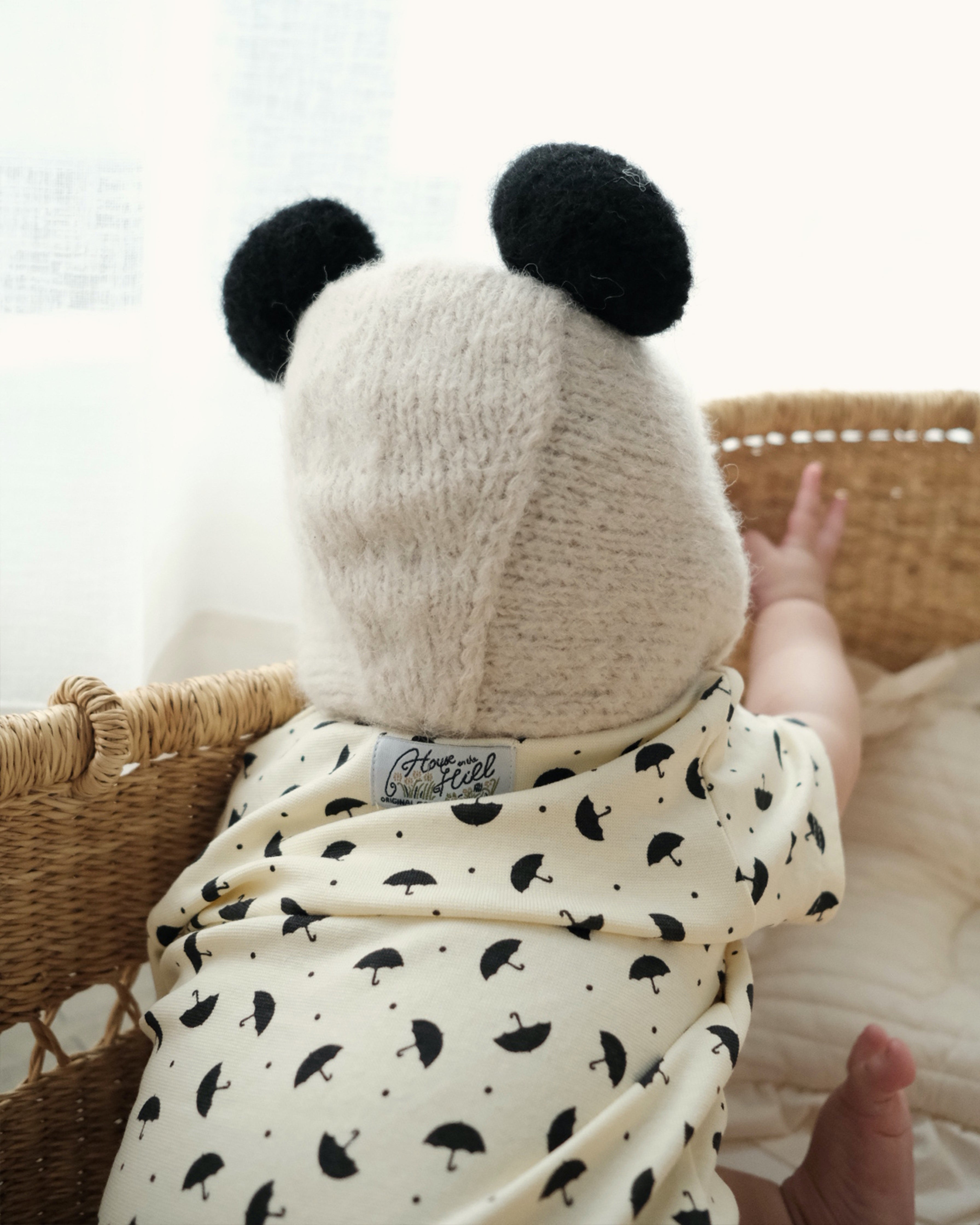 A baby seen from behind wearing a cream knit panda-ear bonnet and a cream onesie with a black umbrella print, sitting in a wicker basket.