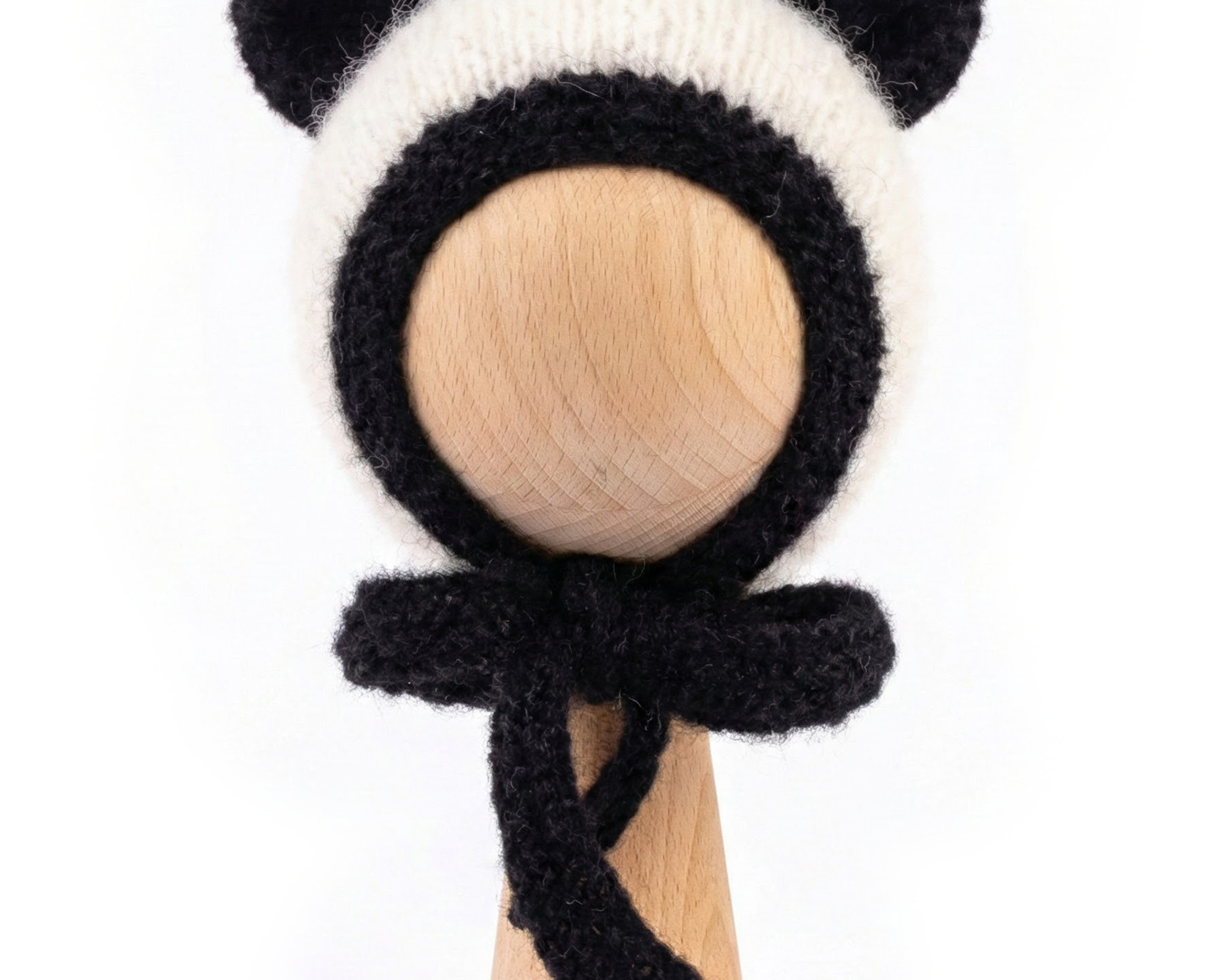 A black and white knitted panda-themed bonnet with ear flaps and a matching bow, displayed on a light wood stand against a white background.