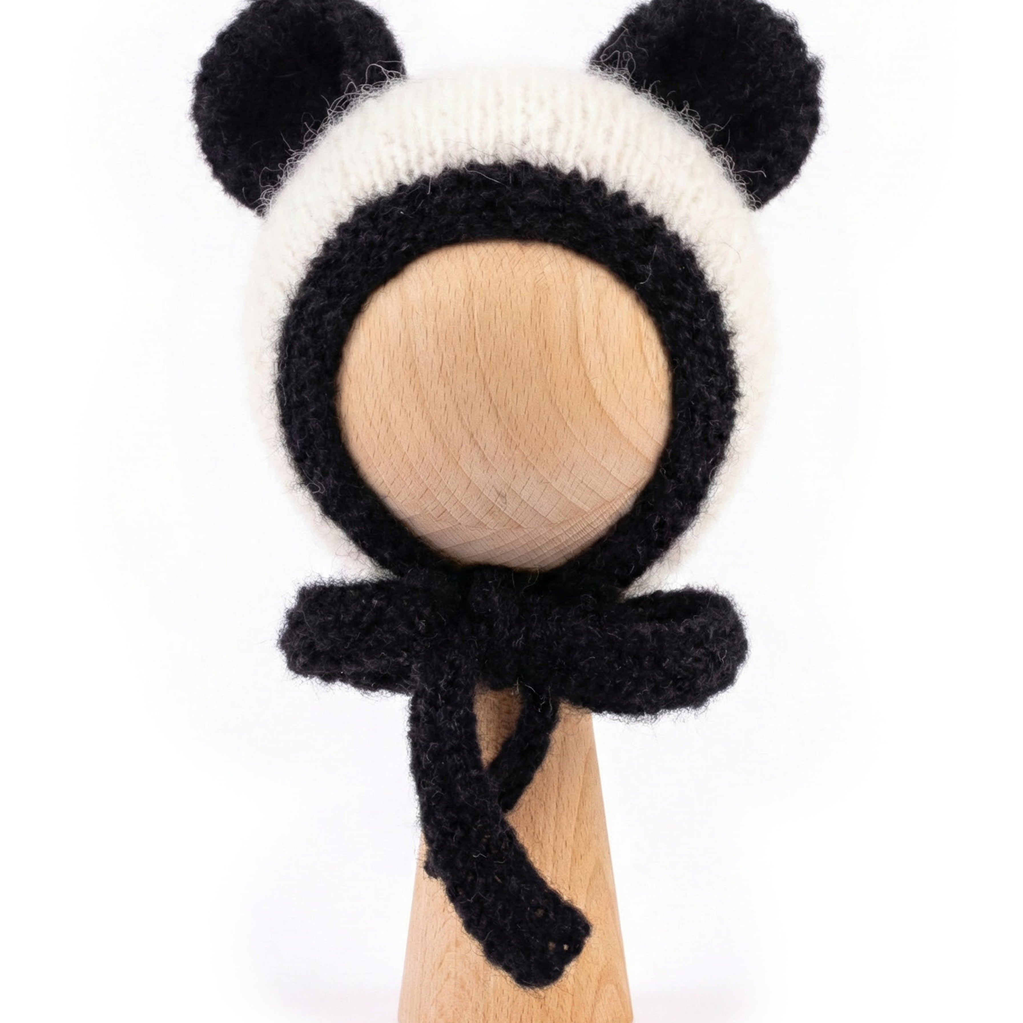 A black and white knitted panda-themed bonnet with ear flaps and a matching bow, displayed on a light wood stand against a white background.
