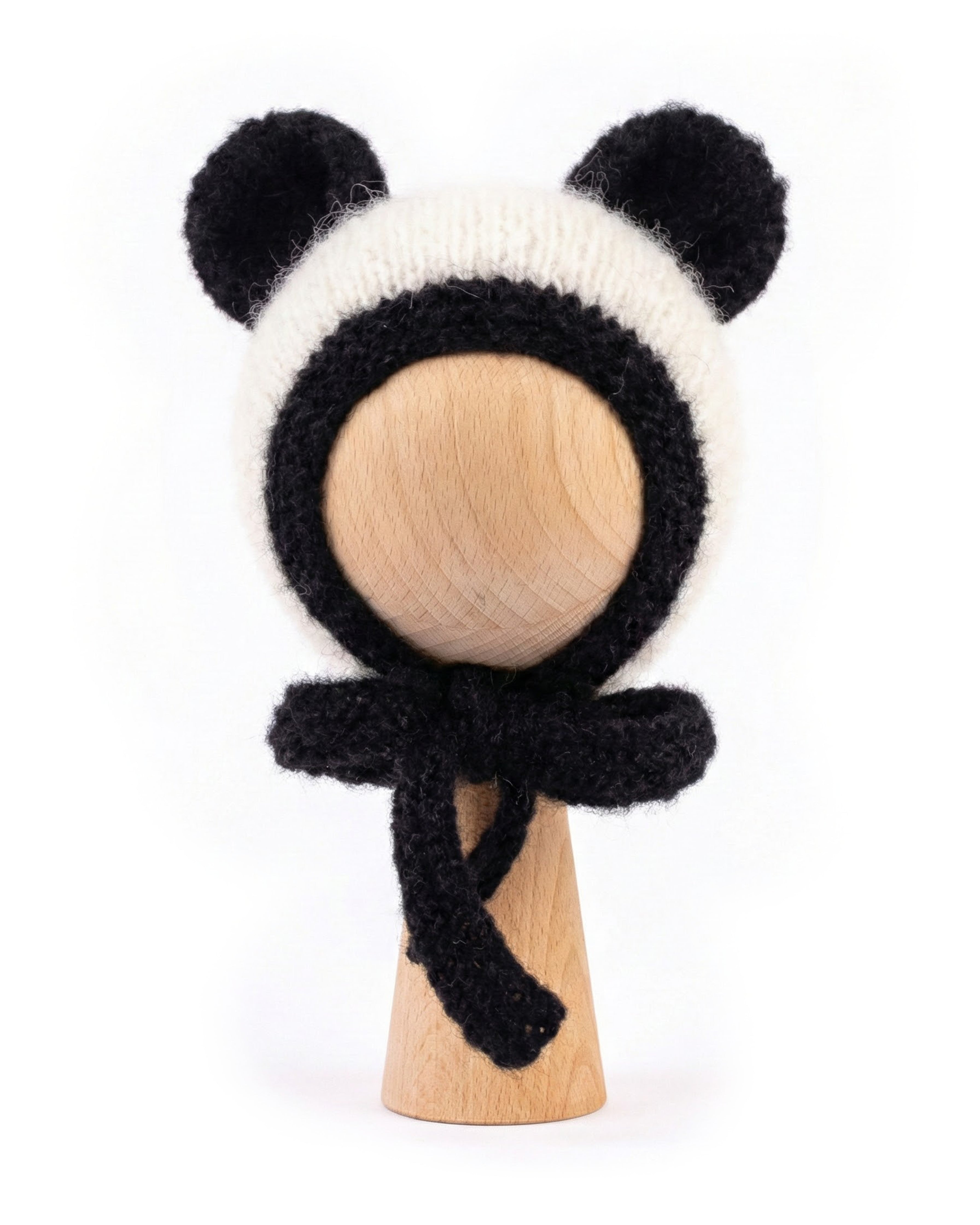 A black and white knitted panda-themed bonnet with ear flaps and a matching bow, displayed on a light wood stand against a white background.