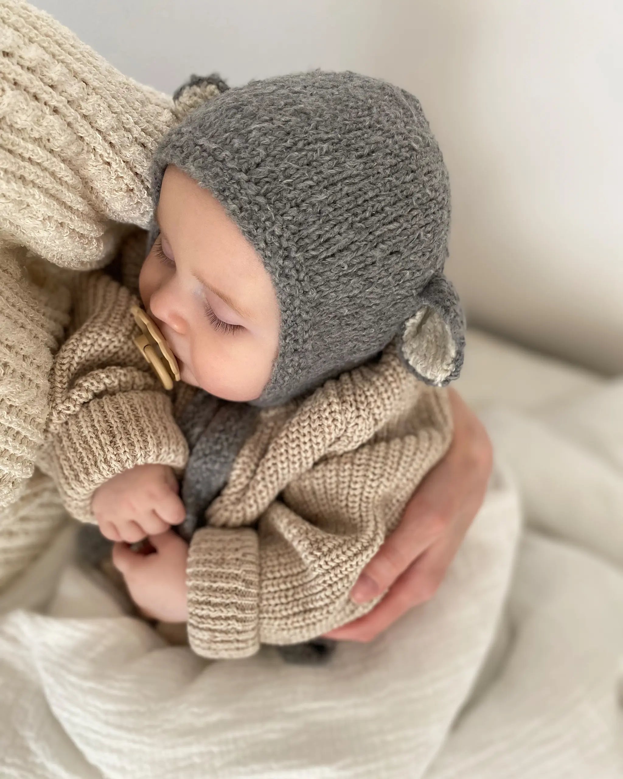 Side view of a baby in the Sheep Bonnet in Charcoal, showing the ecru-lined ear and the knitted tie.