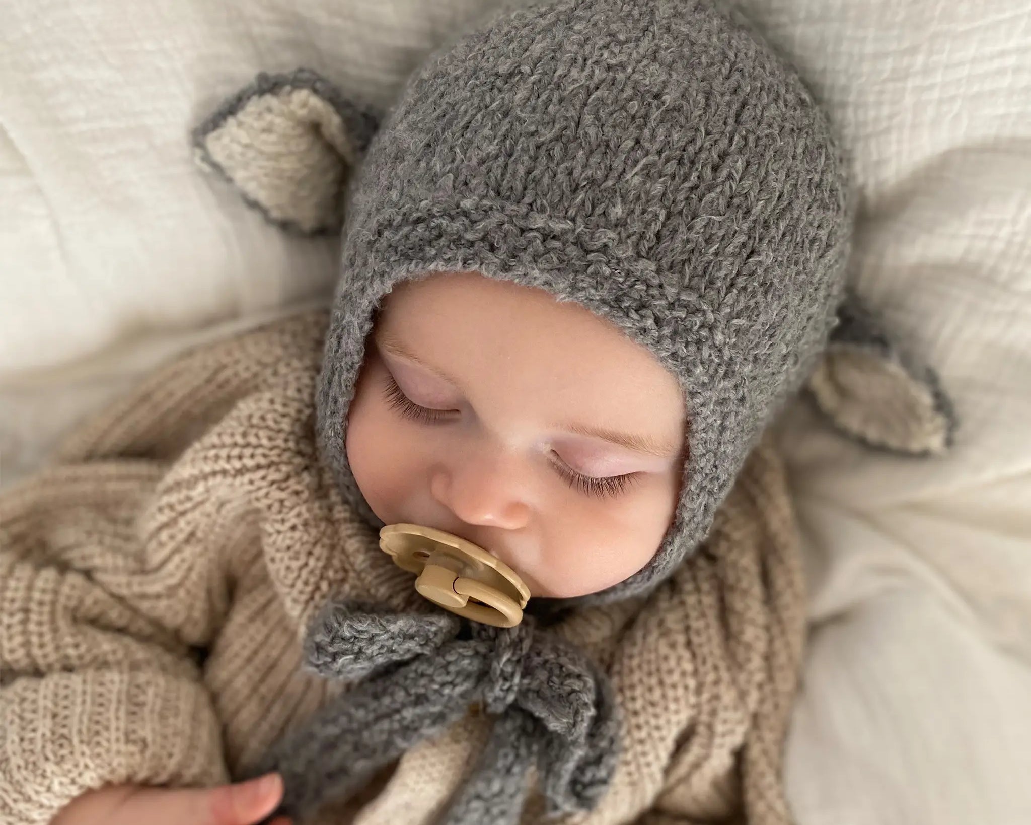 A sleeping baby wearing the Sheep Bonnet in Charcoal, a knitted cotton bonnet with ears and ties fastened under the chin.