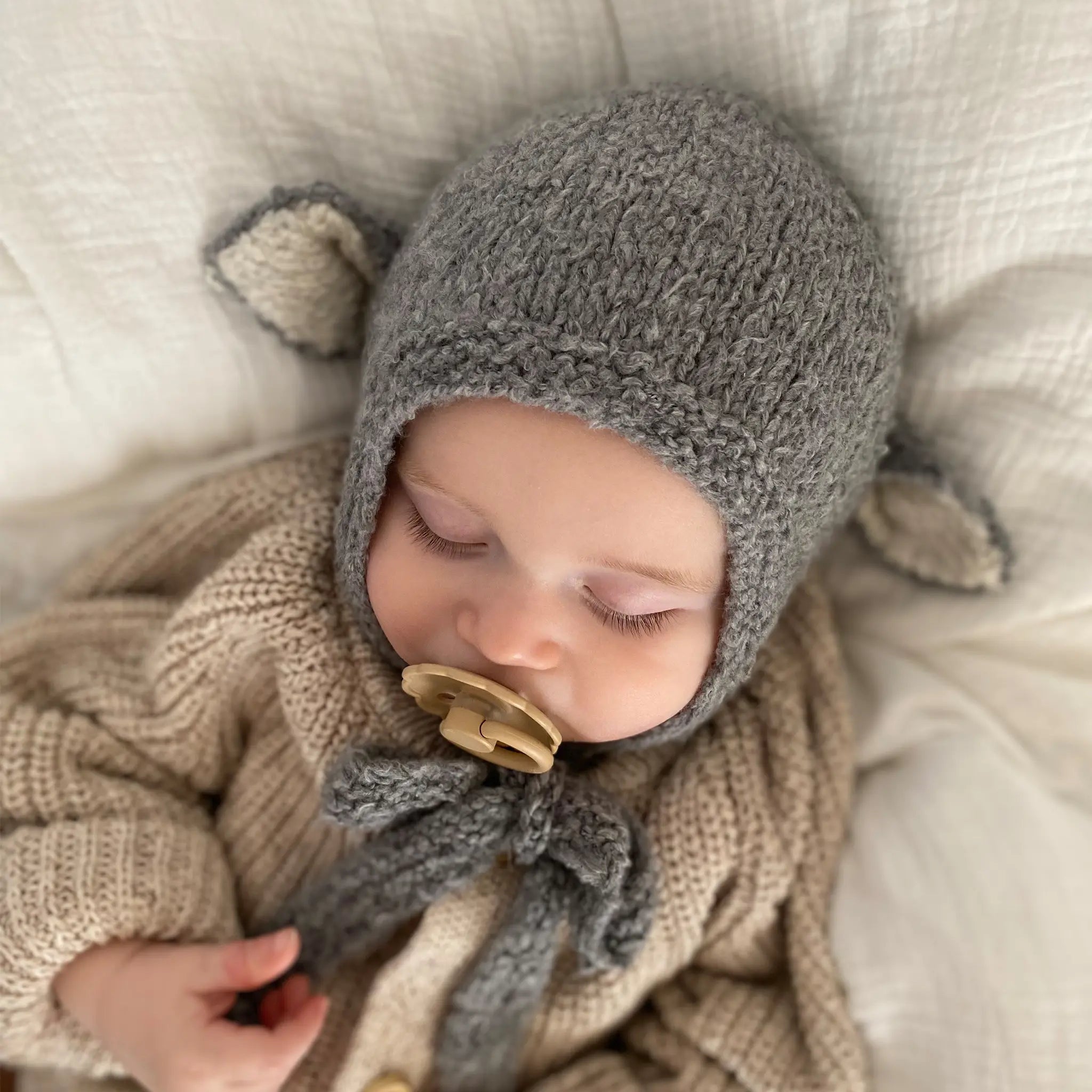 A sleeping baby wearing the Sheep Bonnet in Charcoal, a knitted cotton bonnet with ears and ties fastened under the chin.