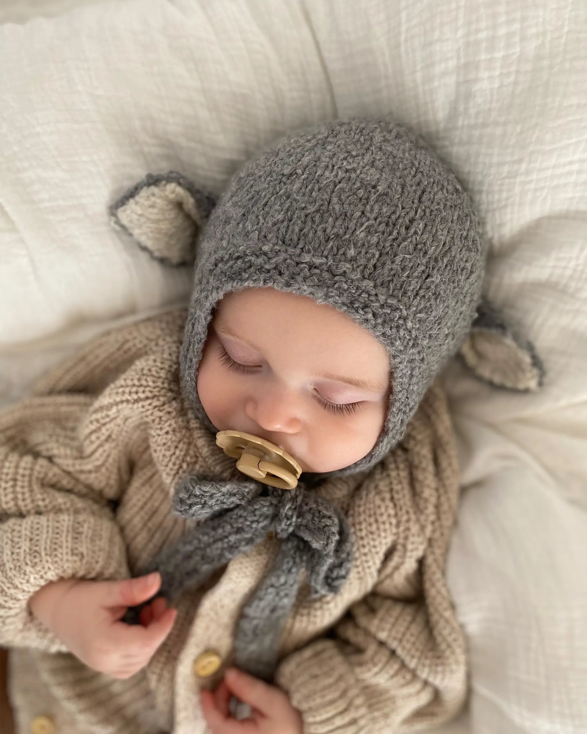 A sleeping baby wearing the Sheep Bonnet in Charcoal, a knitted cotton bonnet with ears and ties fastened under the chin.