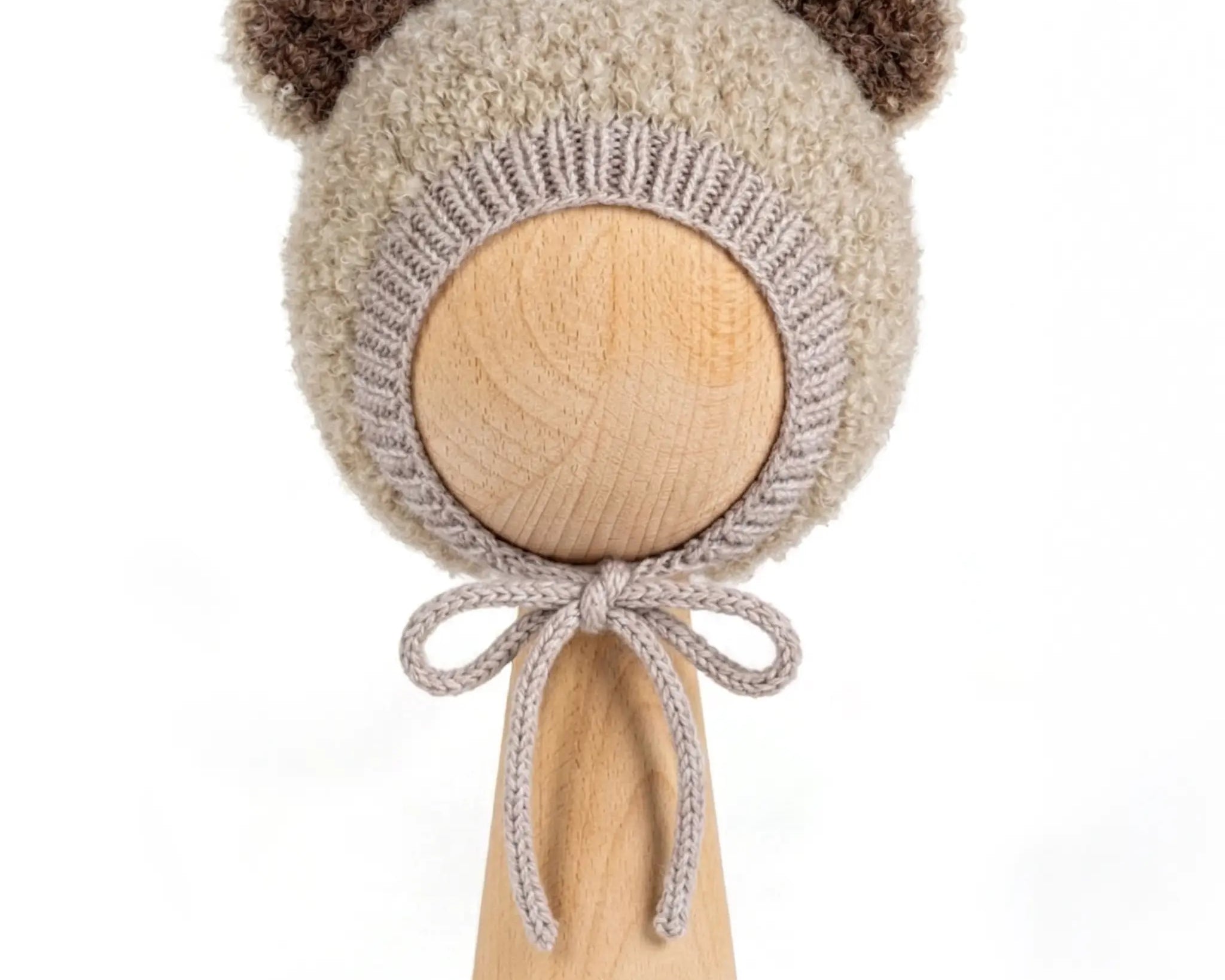 A beige fuzzy knit bonnet with brown pom-pom ears and a matching bow, displayed on a light wood stand against a white background.