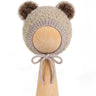 A beige fuzzy knit bonnet with brown pom-pom ears and a matching bow, displayed on a light wood stand against a white background.