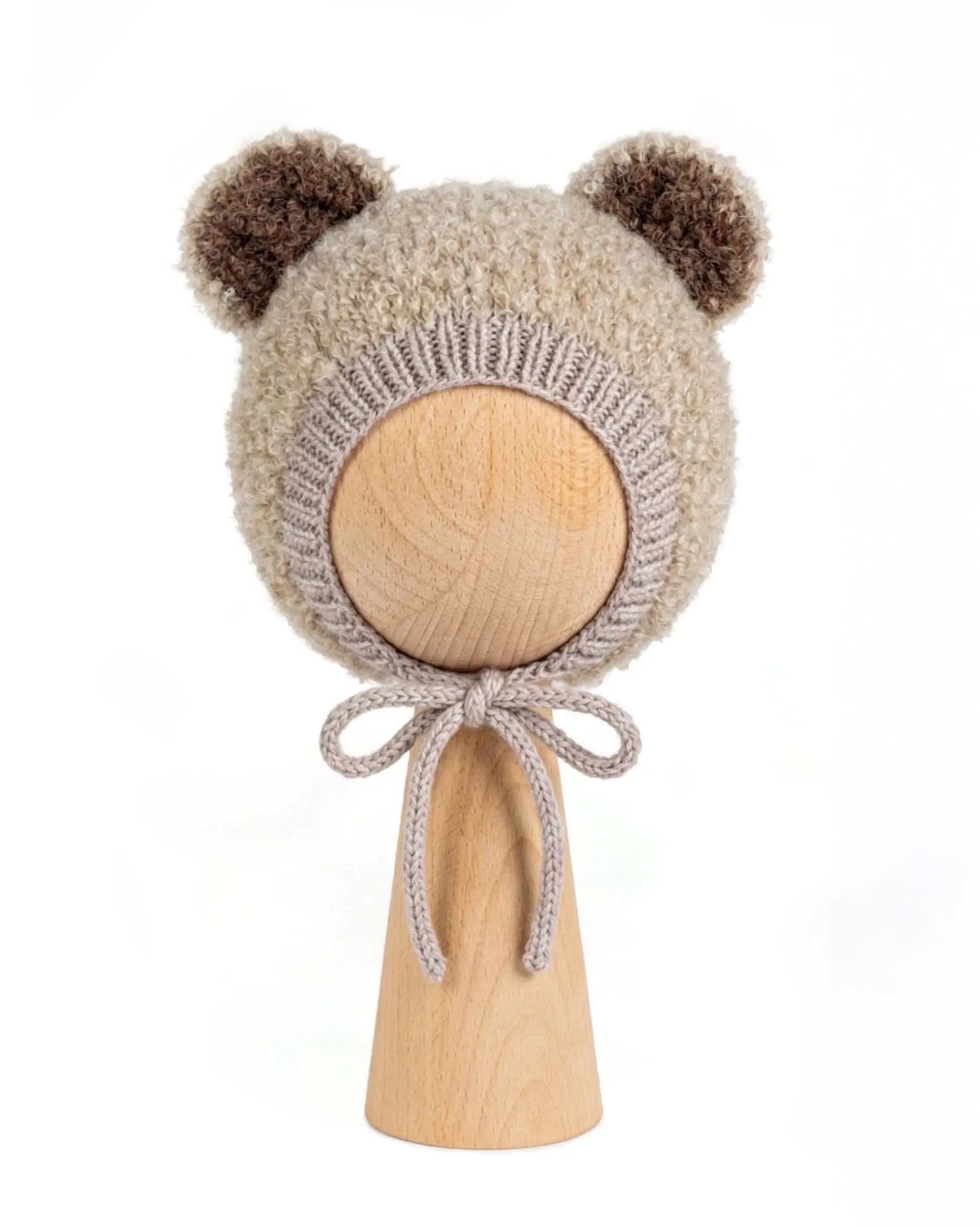 A beige fuzzy knit bonnet with brown pom-pom ears and a matching bow, displayed on a light wood stand against a white background.