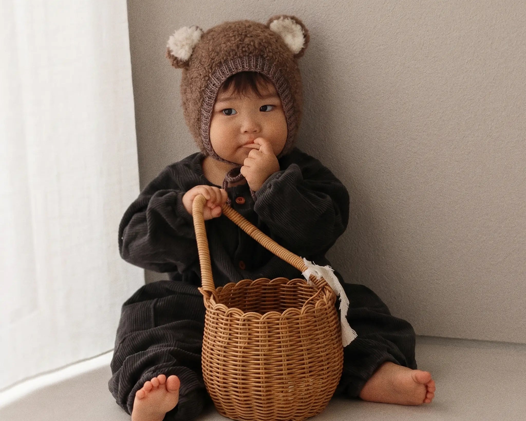 A baby wearing a brown knit bear hat and a black outfit sits on a beige floor next to a window, holding a small woven wicker basket.