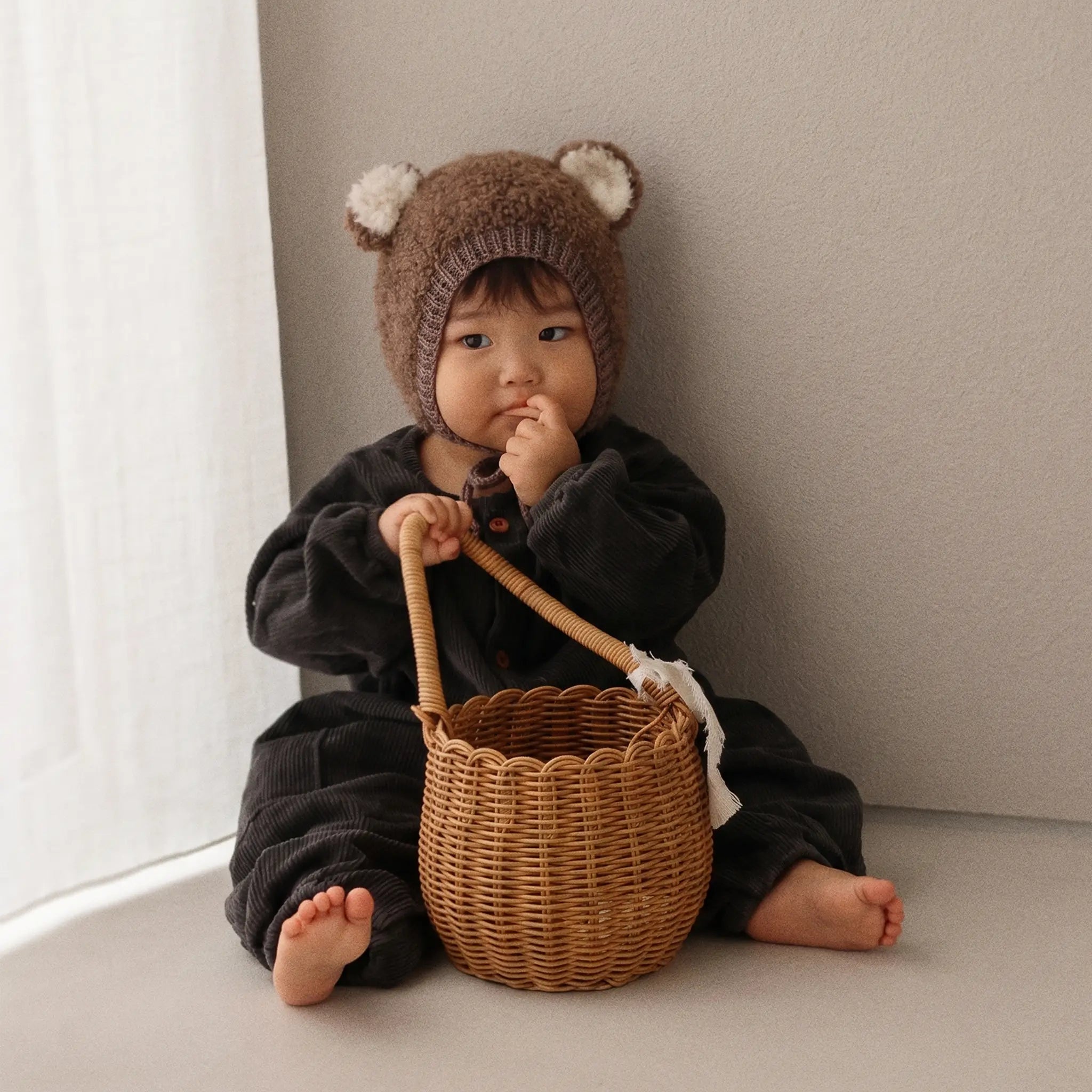 A baby wearing a brown knit bear hat and a black outfit sits on a beige floor next to a window, holding a small woven wicker basket.