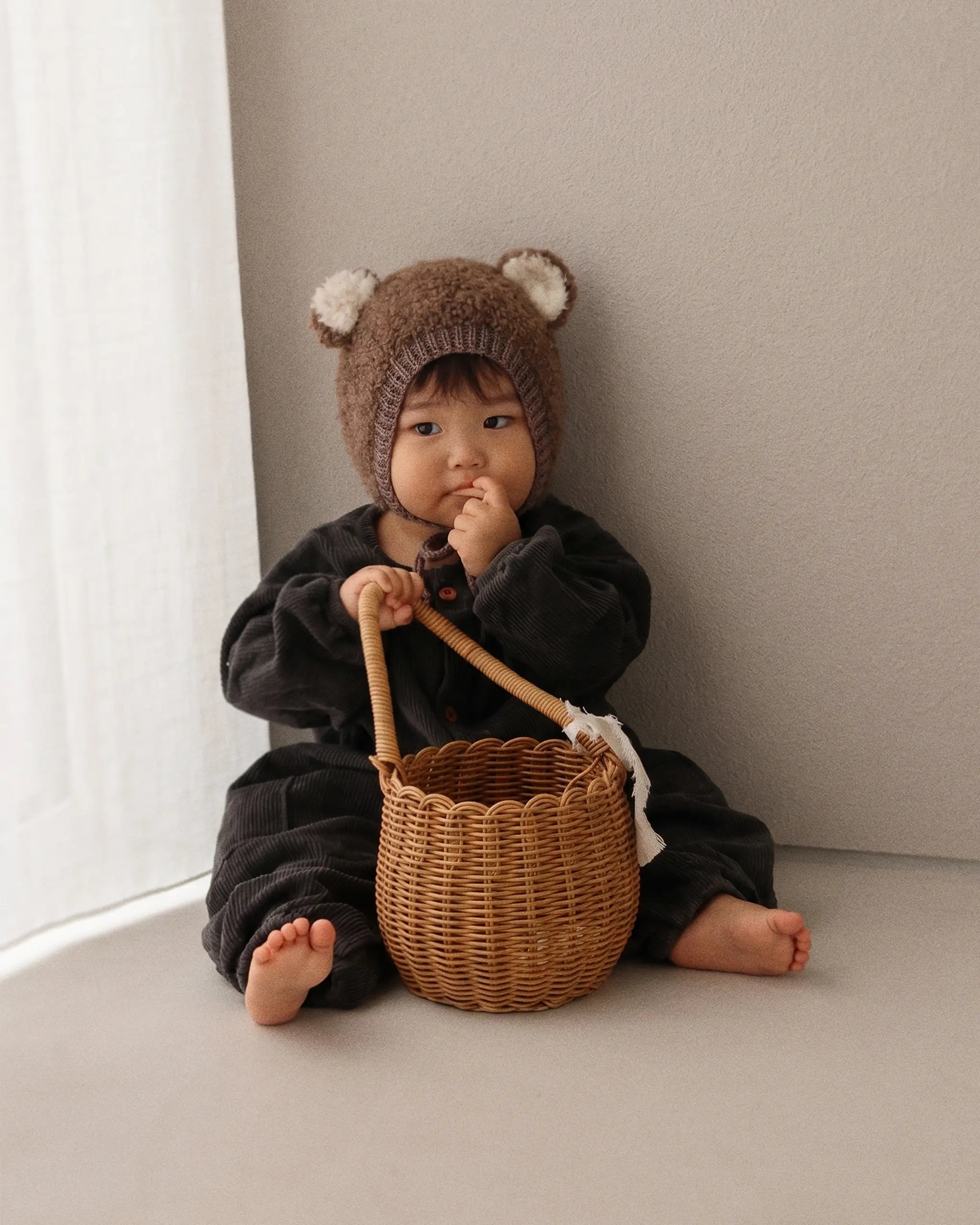 A baby wearing a brown knit bear hat and a black outfit sits on a beige floor next to a window, holding a small woven wicker basket.